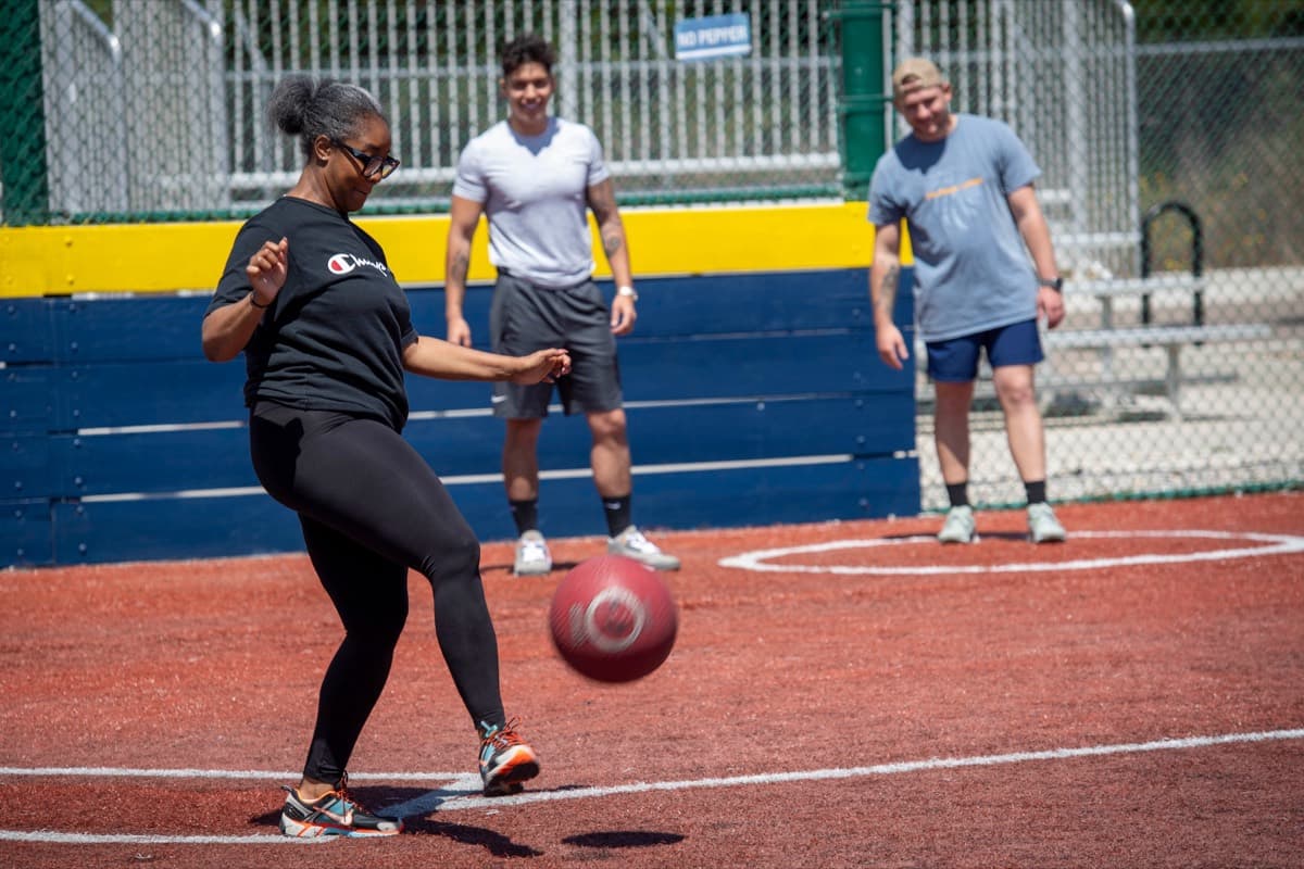 Adult rec league kickball game — a player kicks a red rubber ball at home plate